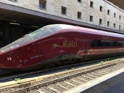 A sleek red high-speed train is stationed on railway tracks at a modern train station, with a beige stone building in the background. The text 'Italo' and 'Smart Cinema' are visible on the side of the train, indicating its branding.