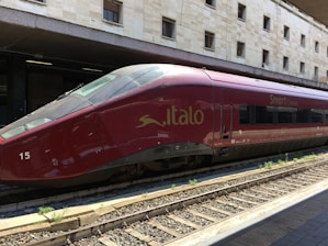 A sleek red high-speed train is stationed on railway tracks at a modern train station, with a beige stone building in the background. The text 'Italo' and 'Smart Cinema' are visible on the side of the train, indicating its branding.