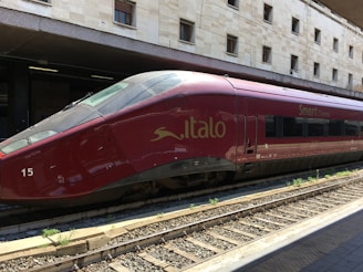 A sleek red high-speed train is stationed on railway tracks at a modern train station, with a beige stone building in the background. The text 'Italo' and 'Smart Cinema' are visible on the side of the train, indicating its branding.