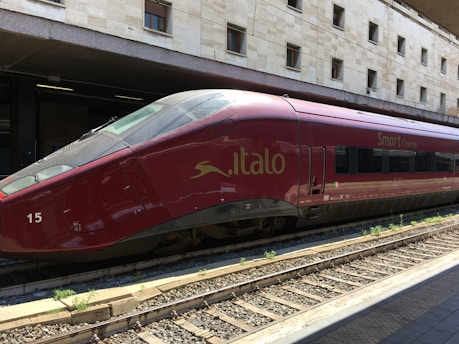 A sleek red high-speed train is stationed on railway tracks at a modern train station, with a beige stone building in the background. The text 'Italo' and 'Smart Cinema' are visible on the side of the train, indicating its branding.