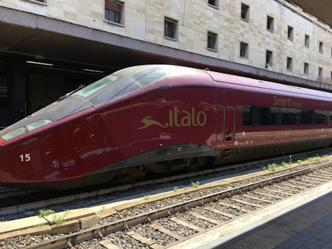 A sleek red high-speed train is stationed on railway tracks at a modern train station, with a beige stone building in the background. The text 'Italo' and 'Smart Cinema' are visible on the side of the train, indicating its branding.