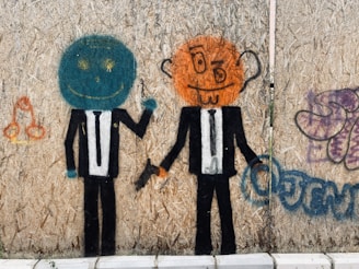 Graffiti art depicting two cartoon figures wearing suits with brightly colored heads. The figure on the left has a turquoise circular head with yellow eyes and a wide smile, while the figure on the right has an orange head with exaggerated facial features, including a crooked smile and prominent ears. Both figures are holding handguns, and the image is painted on a textured plywood surface. Additional graffiti elements include abstract shapes and symbols in different colors surrounding the figures.