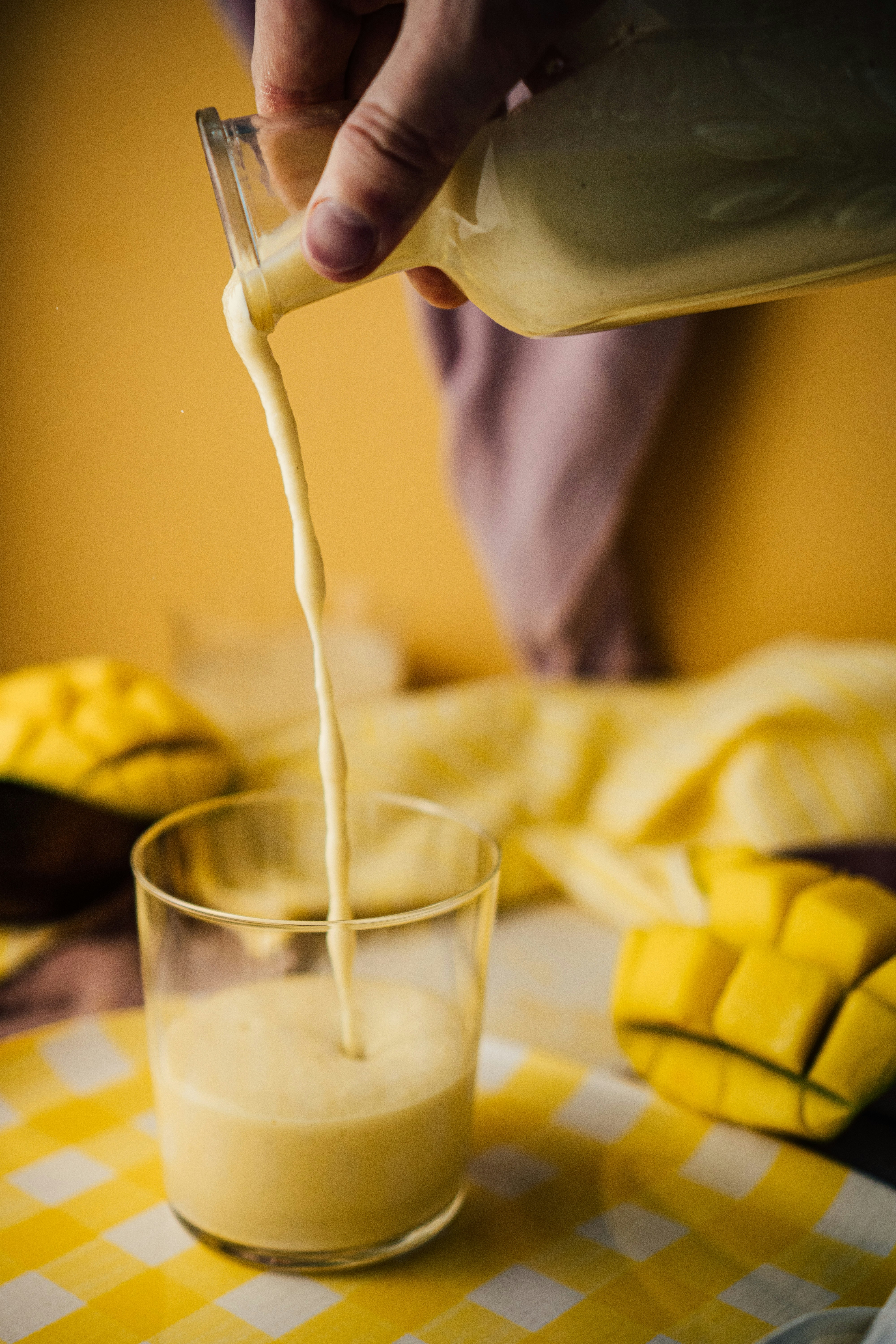 decanter pouring bright orange smoothie mix into tall glass on a table surrounded by cube cut mangoes