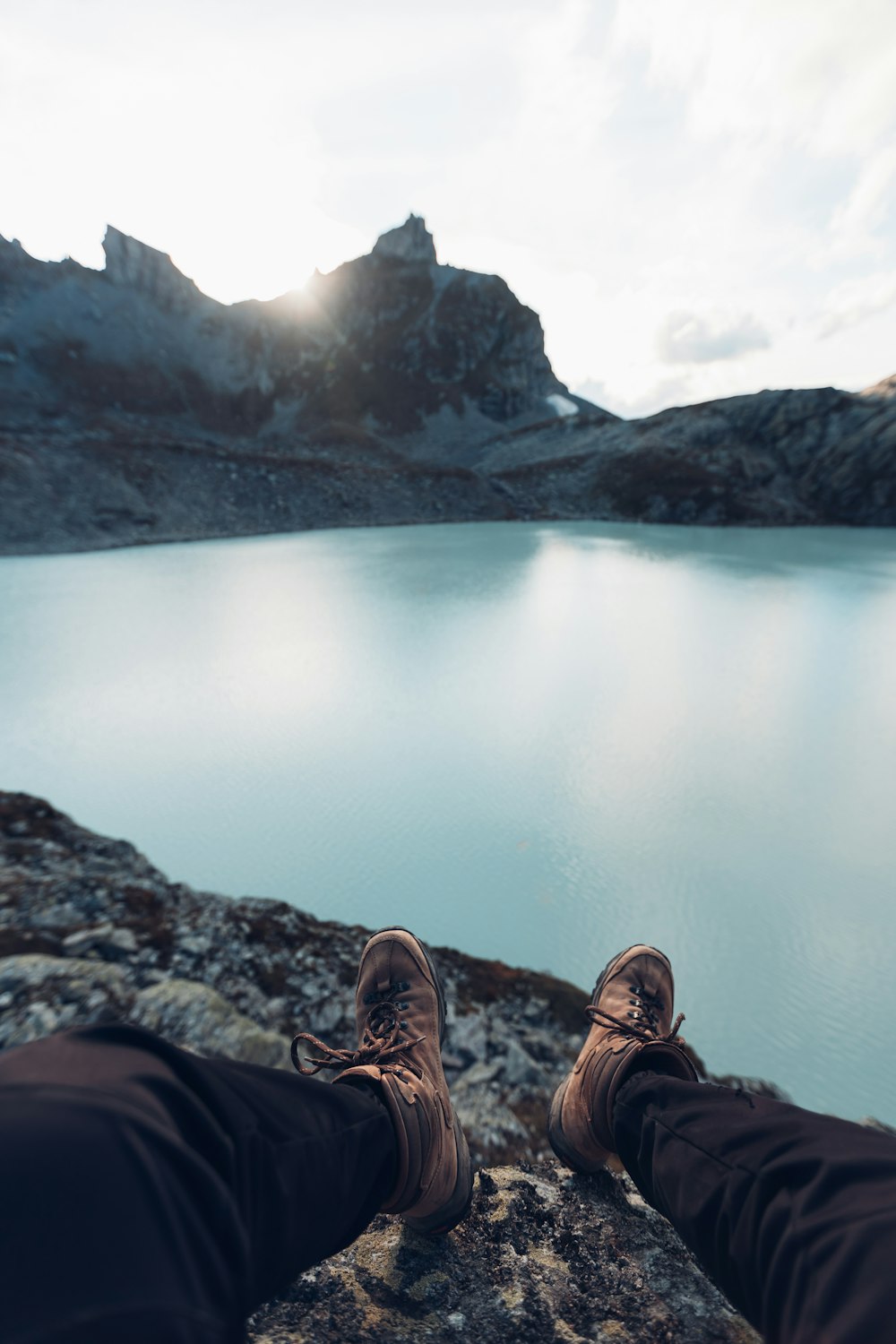 Person in schwarzen Hosen und braunen Wanderschuhen, die tagsüber auf einem Felsen in der Nähe des Sees sitzt