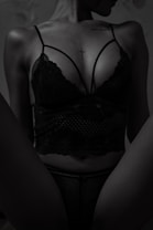 A black and white photograph portrays a person wearing lingerie with a crucifix tattoo on their chest. The lighting creates dramatic shadows highlighting the intricate lace details of the clothing and the contours of the body.