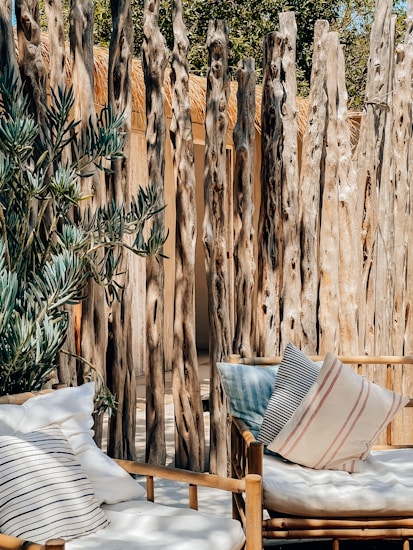 A cozy outdoor seating area with a wooden rustic fence made of vertical logs. The seating features white cushions with a couple of decorative striped pillows. To the left, there is a leafy green plant adding a natural touch to the setting. The sunlight casts soft shadows, creating a warm and inviting atmosphere.