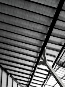 A detailed view of a metallic roof structure showing beams and panels in a geometric arrangement. The panels appear to be made of a translucent material, allowing some light to pass through. A large, abstract sculpture or support structure is visible in the foreground, extending upwards.