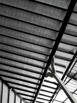 A detailed view of a metallic roof structure showing beams and panels in a geometric arrangement. The panels appear to be made of a translucent material, allowing some light to pass through. A large, abstract sculpture or support structure is visible in the foreground, extending upwards.