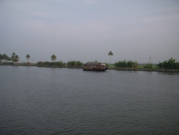 A serene waterway with a traditional houseboat floating calmly in the center, bordered by lush greenery and palm trees. The sky is overcast, adding a touch of tranquility to the scene.