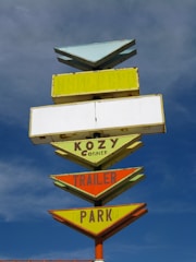 A vintage multi-layered sign with arrow designs, featuring text like 'KOZY Corner TRAILER PARK' against a backdrop of a partly cloudy blue sky. The sign is painted in a mix of pastel and bright colors including yellow, orange, and green, and appears aged.