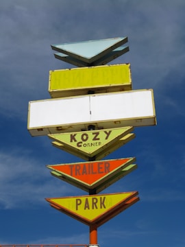 A vintage multi-layered sign with arrow designs, featuring text like 'KOZY Corner TRAILER PARK' against a backdrop of a partly cloudy blue sky. The sign is painted in a mix of pastel and bright colors including yellow, orange, and green, and appears aged.