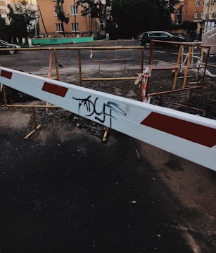 A construction or blockade area on a paved road with a white and red barrier placed in the foreground. The barrier has graffiti on it. Behind the barrier, there are makeshift wooden railings with some caution tape, indicating a restricted or work zone. Residential buildings and some parked cars are visible in the background.