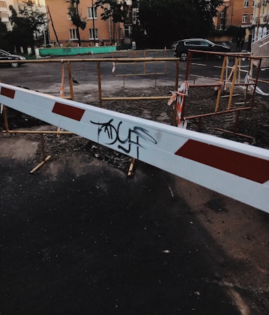 A construction or blockade area on a paved road with a white and red barrier placed in the foreground. The barrier has graffiti on it. Behind the barrier, there are makeshift wooden railings with some caution tape, indicating a restricted or work zone. Residential buildings and some parked cars are visible in the background.