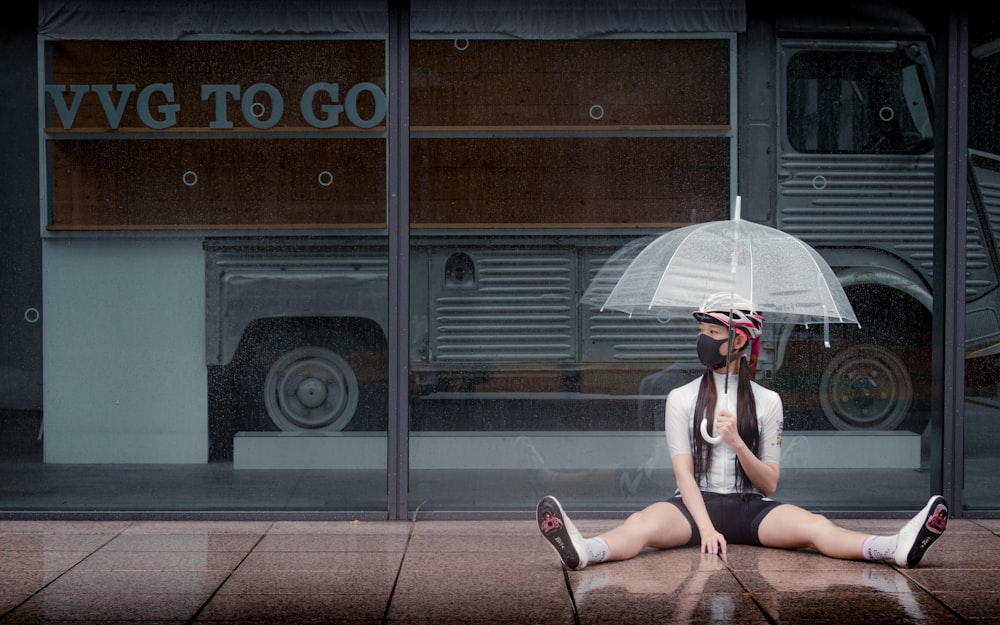 Mujer en camiseta blanca sin mangas sentada en la acera sosteniendo paraguas