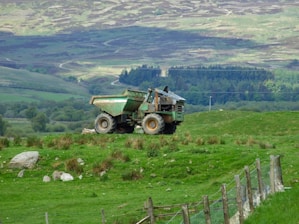 A rugged landscape with a green open field in the foreground, where a large dump truck sits atop a grassy hill. The background features rolling hills and dense forested areas, with a patchwork of green and brown hues. A wooden fence runs along the right side of the image, leading the eye toward the expansive terrain.