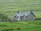 A rustic stone cottage with blue trims is surrounded by lush green fields. The cottage has a pitched roof with chimneys, and there is a stone wall and a wooden gate nearby. Trees dot the landscape in the background, and the scene suggests a serene countryside setting.