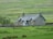 A rustic stone cottage with blue trims is surrounded by lush green fields. The cottage has a pitched roof with chimneys, and there is a stone wall and a wooden gate nearby. Trees dot the landscape in the background, and the scene suggests a serene countryside setting.