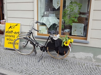 A vintage black bicycle is parked on a cobblestone sidewalk in front of a shop window. The bicycle has a bundle of bright yellow sunflowers on the back. A bright yellow sign with black text advertising a fabric store is standing nearby. The shop window displays various textiles and decorative items, with a leafy plant adding a touch of greenery.