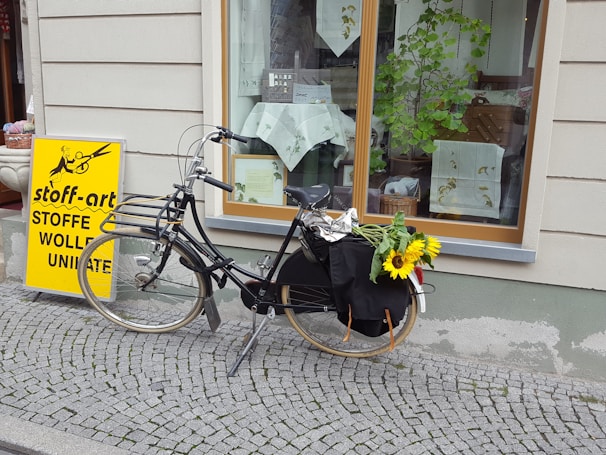 A vintage black bicycle is parked on a cobblestone sidewalk in front of a shop window. The bicycle has a bundle of bright yellow sunflowers on the back. A bright yellow sign with black text advertising a fabric store is standing nearby. The shop window displays various textiles and decorative items, with a leafy plant adding a touch of greenery.