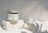 A jar labeled 'Mascarilla Natural Arcilla Verde' sits on two white stone blocks. Next to it, there is a small clear glass bowl with a white spatula-like tool inside. The setting is minimalistic with soft lighting casting gentle shadows, creating a serene and clean ambience.