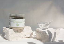 A jar labeled 'Mascarilla Natural Arcilla Verde' sits on two white stone blocks. Next to it, there is a small clear glass bowl with a white spatula-like tool inside. The setting is minimalistic with soft lighting casting gentle shadows, creating a serene and clean ambience.