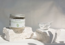 A jar labeled 'Mascarilla Natural Arcilla Verde' sits on two white stone blocks. Next to it, there is a small clear glass bowl with a white spatula-like tool inside. The setting is minimalistic with soft lighting casting gentle shadows, creating a serene and clean ambience.