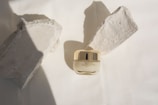 A small jar of Adore Advanced Firming Eye Cream is centrally placed on a neutral background, flanked by two rough-textured stone pieces casting shadows. The jar has a glossy golden cap, contrasting with the matte texture of the surrounding stones.
