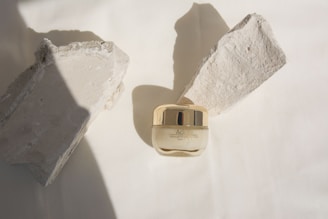 A small jar of Adore Advanced Firming Eye Cream is centrally placed on a neutral background, flanked by two rough-textured stone pieces casting shadows. The jar has a glossy golden cap, contrasting with the matte texture of the surrounding stones.