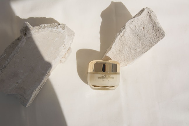 A small jar of Adore Advanced Firming Eye Cream is centrally placed on a neutral background, flanked by two rough-textured stone pieces casting shadows. The jar has a glossy golden cap, contrasting with the matte texture of the surrounding stones.