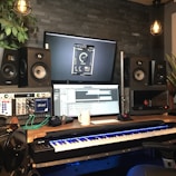 A music production studio setup featuring two large speakers, two computer monitors displaying audio software, a keyboard with illuminated keys, and various audio equipment. The room has a dark brick wall, potted plants, and hanging industrial-style light fixtures providing a cozy atmosphere.