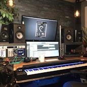 A music production studio setup featuring two large speakers, two computer monitors displaying audio software, a keyboard with illuminated keys, and various audio equipment. The room has a dark brick wall, potted plants, and hanging industrial-style light fixtures providing a cozy atmosphere.