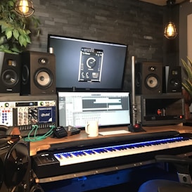 A music production studio setup featuring two large speakers, two computer monitors displaying audio software, a keyboard with illuminated keys, and various audio equipment. The room has a dark brick wall, potted plants, and hanging industrial-style light fixtures providing a cozy atmosphere.