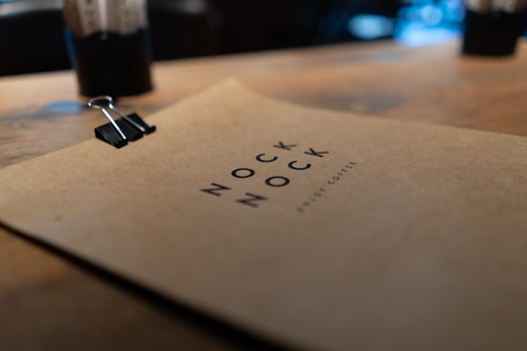 A close-up view of a clipboard with a brown sheet of paper clipped onto it, featuring the text 'NOCK NOCK' and 'ABOUT COFFEE' in a minimalist font. The background is softly blurred, suggesting an indoor setting with a warm ambiance.