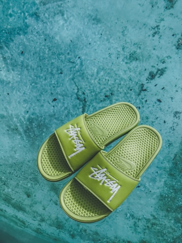 A pair of green slides with white text design rests on a textured concrete surface. The slides have a perforated texture on the footbed and prominently display a brand logo across the straps.