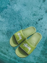 A pair of green slides with white text design rests on a textured concrete surface. The slides have a perforated texture on the footbed and prominently display a brand logo across the straps.