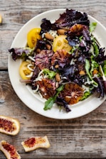 A vibrant salad featuring blood orange slices, yellow cherry tomatoes, almonds, and mixed greens, topped with shredded cheese. The salad is presented in a white bowl set on a rustic wooden surface with pieces of blood orange peel nearby.