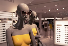 Two mannequins wearing sunglasses and yellow strapless tops are displayed in a well-lit retail store. Shelves filled with various sunglasses are visible in the background, creating a stylish and modern shopping environment.