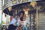 A woman with long curly hair is joyfully embracing a carousel giraffe. The backdrop is adorned with ornate vintage decorations and floral designs. She smiles warmly while holding a face mask in one hand. Sunlight beams through, adding vibrancy to the scene.