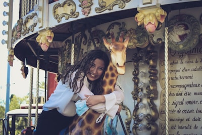 A woman with long curly hair is joyfully embracing a carousel giraffe. The backdrop is adorned with ornate vintage decorations and floral designs. She smiles warmly while holding a face mask in one hand. Sunlight beams through, adding vibrancy to the scene.