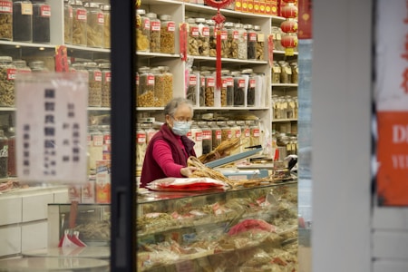 An elderly person wearing a face mask and a red jacket, standing inside a shop with shelves lined with numerous jars containing various herbs and dried goods. The shop has a traditional appearance, with red and gold decorations hanging from the shelves.