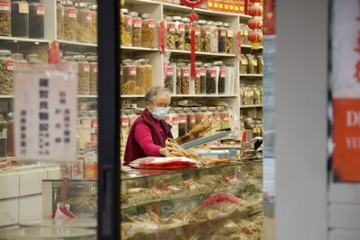 An elderly person wearing a face mask and a red jacket, standing inside a shop with shelves lined with numerous jars containing various herbs and dried goods. The shop has a traditional appearance, with red and gold decorations hanging from the shelves.