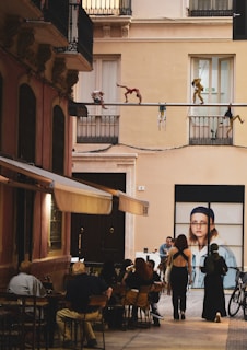 A narrow street features outdoor seating where people are gathered at tables under awnings. The backdrop includes a large advertisement with a woman in casual clothing and smaller mannequin-like figures posed along a pipe above. The architecture suggests a European setting, with ornate balconies and warm-colored buildings.