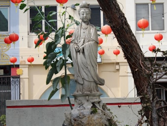 A stone statue of a serene figure stands in a garden setting, surrounded by lush green foliage. In the background, a building facade is adorned with multiple red lanterns that hang in orderly rows, adding a festive and cultural ambiance. A large tree trunk and branches partially frame the scene, enhancing the outdoor environment.