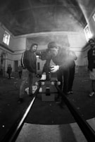 A group of people are in an industrial or abandoned space with high ceilings, looking intently at a camera mounted on a slider. They seem to be preparing for a shoot, with the main focus on one individual adjusting the camera. Smoke, possibly from a smoke machine, adds a hazy atmosphere to the scene.