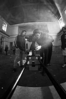 A group of people are in an industrial or abandoned space with high ceilings, looking intently at a camera mounted on a slider. They seem to be preparing for a shoot, with the main focus on one individual adjusting the camera. Smoke, possibly from a smoke machine, adds a hazy atmosphere to the scene.