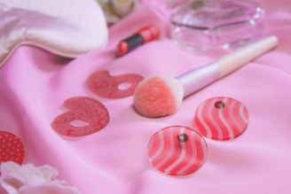 A soft pink background with various beauty items arranged on it, including a makeup brush with a fluffy pink tip, a small tube of lipstick, two round decorative plastic pieces with striped designs, and a clear, round bottle. The scene is set with a gentle, feminine aesthetic.