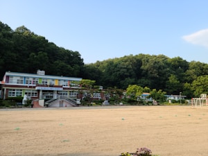 A colorful school building with a playground in front, surrounded by lush green trees. The structure is a single-story building with a variety of bright colors on its facade. The playground area appears to be sandy, and there are goalposts set up for sports activities. The background consists of a dense forest under a clear blue sky.