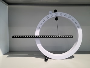 A modern, circular perpetual calendar with a minimalist design is displayed. It consists of a white ring marked with months and a horizontal black bar marked with days. Small black and white spheres are used to indicate the current month and day. The calendar is set against a neutral background, casting soft shadows.