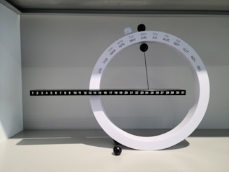 A modern, circular perpetual calendar with a minimalist design is displayed. It consists of a white ring marked with months and a horizontal black bar marked with days. Small black and white spheres are used to indicate the current month and day. The calendar is set against a neutral background, casting soft shadows.
