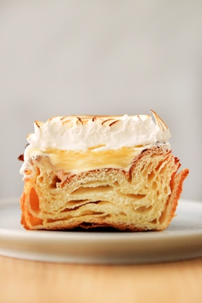 A close-up of a slice of pastry showing its flaky, layered texture. The top is topped with a generous amount of cream, slightly browned, indicating a toasted or baked finish. The pastry sits elegantly on a simple white plate with a blurred background.